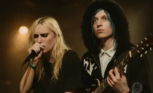 Two musicians perform on stage: a woman sings into a microphone, and a person with blue hair plays a guitar.