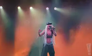 A woman with long blonde hair, wearing a black cap and outfit, sings into a microphone on a stage with orange lighting and smoke effects.