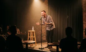 A man with tattoos on his arms holds a microphone while wearing a white t-shirt on a blue stage.