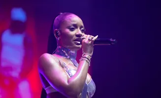 A woman sings into a microphone on a stage, wearing a sparkly silver top and choker.