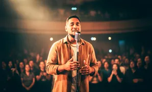 A man sings into a vintage microphone on a dimly lit stage, with an audience blurred in the background.