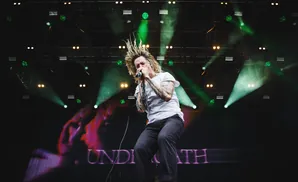 A male singer with wild blonde hair sings into a microphone on a stage with green lights and smoke.