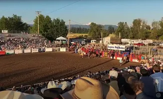 Rodeo arena with bull riders, horses, and spectators in bleachers under a blue sky.
