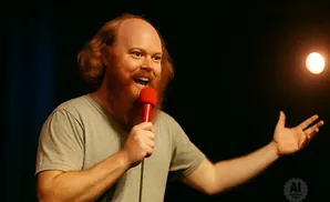 A bald man with a red beard laughs into a red microphone on stage.