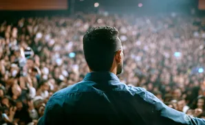 Man in blue shirt on stage addresses a large, blurred audience under bright lights.
