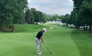Golfer in blue shirt and grey pants tees off on a green golf course with trees and grandstands.