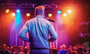 Comedian on stage with a microphone in front of a cheering audience, illuminated by colorful stage lights.