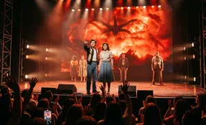 A stage musical performance with actors in period costumes in front of a dramatic screen projection, with an audience in the foreground.