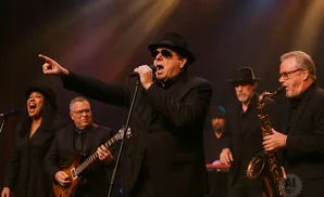 A band performs on stage, with a lead singer in a black suit and hat singing into a microphone.