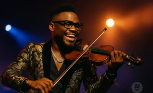 A Black man in a patterned jacket plays the violin with a broad smile, under warm stage lights.