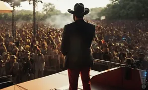 Man in cowboy hat and red pants on stage facing a large cheering crowd at an outdoor concert.