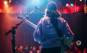 A musician with a guitar is on stage, facing away from the camera, bathed in colorful stage lights.