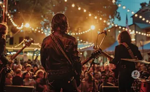 A rock band performs on an outdoor stage at dusk, lit by string lights, with a crowd in the background.