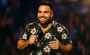 A smiling man in a floral shirt holds a microphone on stage.