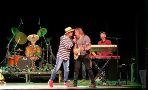 Two men sing and play guitar on stage, with a drummer and keyboardist behind them.
