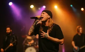 A male singer with long blonde hair and tattoos on his arm performs on stage, illuminated by colorful spotlights.