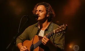 Musician with curly hair singing and playing an acoustic guitar on stage.