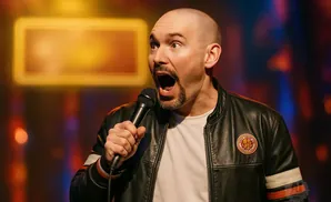 A bald comedian with a goatee holds a microphone and speaks into it with his mouth open in surprise.