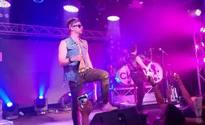 Singer in a denim vest with stars and an American flag bandana performs on stage with bandmates.