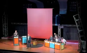 A debate stage with five people on it and two podiums, a large pink screen behind them.