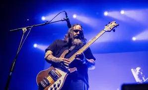 A long-haired, bearded man plays a bass guitar on a stage with blue lighting.