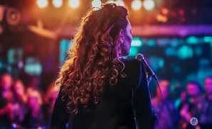 A woman with curly red hair sings into a microphone on a dimly lit stage with colorful lights.