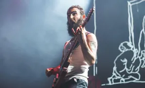 Musician with a beard plays guitar on stage, illuminated by spotlights.