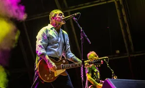 A tattooed male singer plays a guitar on stage with purple and yellow stage lighting.