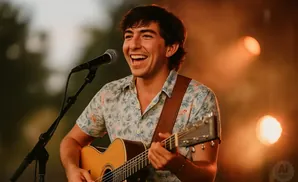 A man in a floral shirt happily plays guitar and sings into a microphone on a stage with warm lighting.