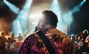 Singer with curly hair in a floral shirt on stage, with a crowd and stage lights behind.