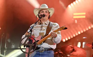 A man in a cowboy hat and striped shirt plays a guitar on stage with warm lighting.