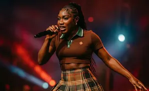 A woman with braided hair performs into a microphone on stage, wearing a brown cropped polo and a plaid skirt.