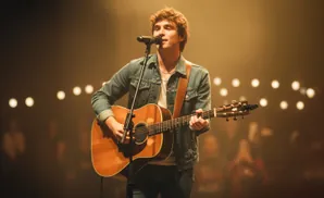 A young man in a denim jacket sings and plays an acoustic guitar on stage.