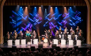 A jazz band performs on a brightly lit stage with a patterned blue and purple backdrop.