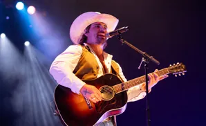 Musician in a cowboy hat sings and plays an acoustic guitar on a dimly lit stage with spotlights.