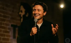 A man with dark wavy hair smiles broadly while holding a microphone, gesturing with his hands on a dimly lit stage.