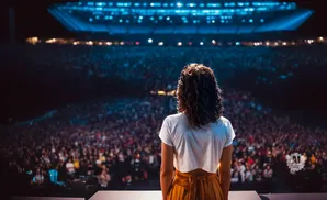 A woman in a white shirt and orange skirt stands on stage facing a large, dimly lit audience at a concert.