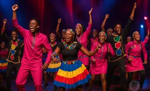 A group of young people in vibrant, colorful traditional African attire singing and dancing energetically on stage.