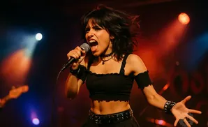 A woman with dark, messy hair sings into a microphone on a dimly lit stage, illuminated by red and blue spotlights.