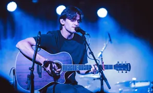 A male musician plays an acoustic guitar on a dimly lit stage, bathed in blue light.