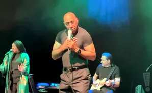 A man in a black t-shirt sings into a microphone on stage, with two musicians beside him.
