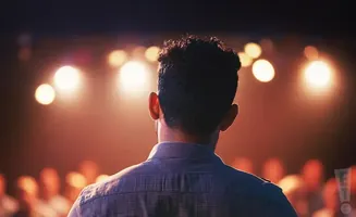 A man's back, facing a stage with bright lights and an audience.