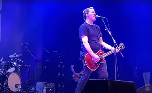 A man in a black t-shirt plays a red electric guitar and sings into a microphone on a stage with blue lighting.