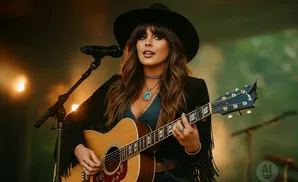 Woman in a black hat and fringed jacket plays an acoustic guitar on stage.