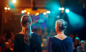 Two women stand facing away from the camera on a dimly lit stage, bathed in colorful spotlights.