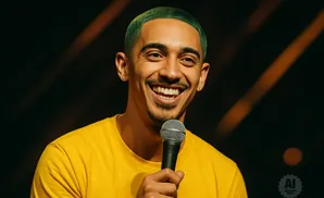 A man with green hair smiles widely while holding a microphone and wearing a yellow shirt.