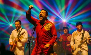 A lead singer in a red jumpsuit belts into a microphone on stage, fists raised, with a band playing behind him under colorful lights.