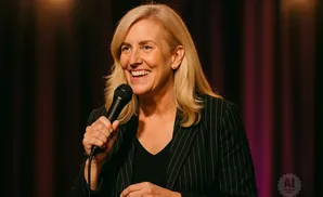 A blonde woman in a pinstripe jacket smiles and holds a microphone on stage.