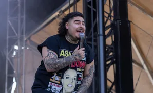A tattooed man with a mullet holds a microphone and smiles onstage in a black t-shirt.