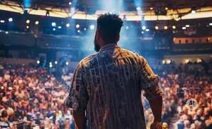 Man in patterned shirt faces a cheering crowd from a stage lit by spotlights.